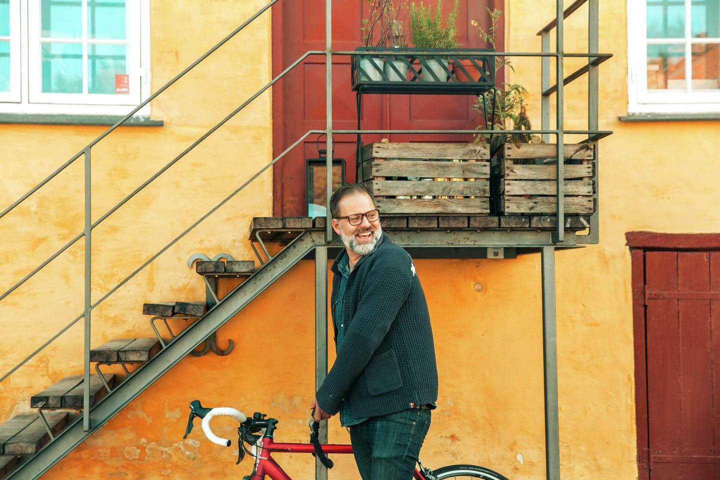 Mand står med cykel foran gult hus og trappe og smiler, fotografi