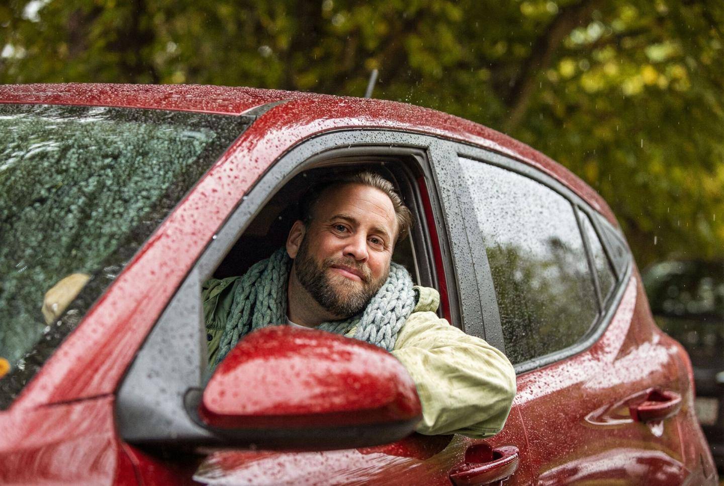 En person sidder i en rød bil med vinduet rullet ned på en regnvejrsdag og hviler armen på døren.